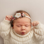 девочка новорожденная лежит в белом свитере на голове цветочек портрет на сером фоне