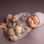 девочка новорожденная спит рядом с мишкой в сереневом вязанном костюме в чепчике эльф