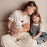 мама с двумя детьми целует старшую дочку девочка смеется новорожденный спит