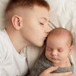 старший брат целует родившегося маленького братика на белом постельном белье